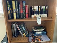 Front view of wooden bookshelf showing two shelves filled with fiction books by John Grisham, Ruth Ware, Dennis Lehane, John Sandford, Jeffrey Deaver, and boxed sets of hardcover books.