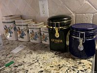 All six canisters shown together on kitchen counter under outlet; four Daeware matching floral ceramic canisters, one black Gevalia coffee canister, one dark blue canister
