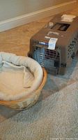 Wide photo showing plastic brown cat carrier with metal door and oval wicker basket cat bed with fleece liner inside, both placed on carpet and tile floor.