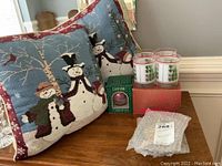 Photo showing two snowman-themed decorative pillows, one new boxed Dansk Christmas ornament, and one set of four Christmas drinking glasses with red rim and green Christmas tree design on a red box.