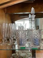 Photo showing crystal champagne flutes, crystal decanter with stopper, and candle holders on shelf, some monogram 'V' visible on flutes.