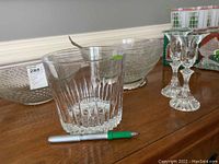 Glass champagne bucket, two glass salad bowls including one with silver-plated rim, and two clear glass candle holders on wood surface with pen for scale.