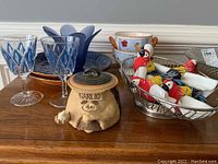 Photo showing blue ceramic plates, blue glass goblets, ceramic soup bowls with rooster design, garlic canister with face, wire basket, and parrot napkin rings with white napkins.