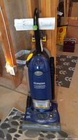 Front view of blue Eureka The Boss SmartVac upright vacuum cleaner standing on a basement floor with manual rolled and attached to handle.
