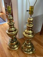 Pair of matching Westwood brass table lamps with ornate rounded designs, no shades attached, cords wrapped around base, standing on hardwood floor.