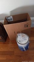 Mostly full gallon of Benjamin Moore white interior paint can with lid, placed near a cardboard box on a hardwood floor.