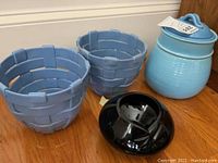 Two woven ceramic lanterns in light blue glaze, blue ceramic ginger jar with lid, and black glass planter on wood floor.