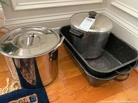 Two stainless steel stock pots, one with black knob lid and black handles, one 8 quart Revere Ware with copper bottom, placed near two black speckled enamel roasting pans stacked.