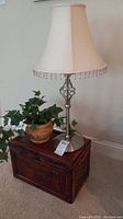Woven rattan box, terra cotta faux plant pot, and silver metal lamp with cloth shade shown together, items placed on carpeted floor against beige wall.