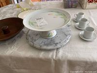 White ceramic pedestal cake plate with hand painted pastel floral design on top and side, resting on round marble Lazy Susan base.