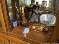Photo shows a gold plate percolator with a black handle, gold plate butter dish with a butter spreader on a wooden surface, and two pewter bread trays placed behind.