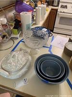 Photo showing all three bowl types: three blue nested ceramic bowls, white china serving bowl with lid, and clear glass footed bowl on kitchen counter.
