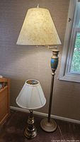 Brass table lamp standing on floor with white pleated lampshade missing finial, and brass swing arm floor lamp with cream paper shade beside window.