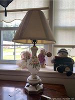 Porcelain table lamp with floral painted base and cloth shade, displayed on windowsill with decorative dolls in background.