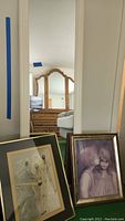 Tall framed wall mirror mounted on a wall reflecting a dresser and bed in the background. Two framed prints leaning against the wall in front of the mirror; one depicts ballet dancers and the other shows a boy and girl.