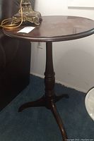 Full view of wooden pie table showing round top, single pedestal, and three splayed legs on dark carpeted floor next to wall.