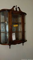 Front view of small wall-mounted wood and glass curio cabinet showing curved glass front and decorative wooden top with finials
