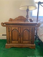 Front view of one wooden nightstand showing two cabinet doors, metal hardware, a missing knob, and a carved decorative back panel.
