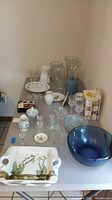 Photo showing assorted glassware items including shot glasses, vases, decanter, plates, ceramic sugar creamer, and Lenox vase arranged on table.