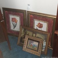 Four framed wall art pieces leaning against a wall on carpet. Two large floral prints with tulips have pink and gold mats. Two smaller ornate gold framed paintings leaning in front.