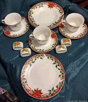 Photo showing multiple plates, cups, saucers, and napkin rings arranged on a blue cloth. The items have poinsettia and holly floral patterns in red, green, and yellow on white ceramic.