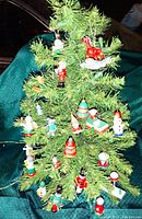 Photo showing assortment of 21 mini wooden handcarved Christmas ornaments hanging on an artificial green Christmas tree display. Various figures including soldiers, animals, drums, and other holiday shapes are present.
