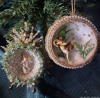 Photo showing two vintage handmade Christmas ornaments hanging on a tree branch: one with a ram figurine and pearl beaded border, and one with an angel figurine surrounded by green beads and pearls.