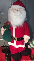 Full front view of 3-1/2 feet tall plush Santa Claus figure dressed in classic red suit and holding green sack of gifts.