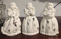 Front view showing three white porcelain angels each holding different objects, with star and snowflake cut out patterns and gold accent designs on gowns.