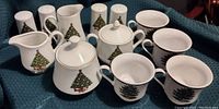 Full set of white ceramic Christmas tree servingware displayed on blue textured cloth including cups, sugar bowl with lid, creamers, and salt and pepper shakers.