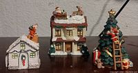 Photo of three mini holiday buildings: white church, two-story house with Santa on roof, and decorated Christmas tree, all snow-covered.
