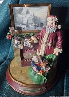 Front and angled views of the Santa Claus figurine showing details of the fireplace, stockings, toys in the green sack, and framed Thomas Kinkade artwork.