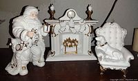 Grouped shot of Santa figurine, porcelain fireplace mantle with stockings, white armchair with sitting dog, all with 22k gold gilded details.