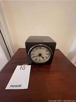 Front view of the Pottery Barn cube clock showing the clock face with Roman numerals and black hour, minute, and second hands. Clock is set on a wooden surface with neutral background.
