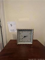 Front view of sterling silver square clock on wood surface with visible rope-style border frame and clock face.