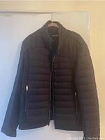 Front view of the black men's Karl Lagerfeld quilted jacket hanging on a hanger. Shows the front zipper and pockets with zipper pulls, and logo patch on left sleeve.