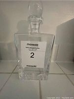 Front view of square clear glass liquor decanter with stopper on tiled surface.