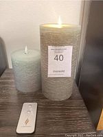 Lot of two Luminar pillar candles in different sizes with LED flame tops and a Luminar branded remote control, set on wooden surface.