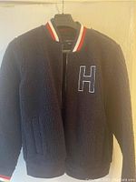 Front view of the black fleece jacket hanging on a black hanger, showing the textured material and embroidered 'H' on the chest.