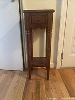 Front view of small wooden table showing single drawer, four legs with turned detail, and lower shelf. Table is medium brown stained wood.