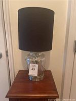Front view of the lamp showing black shade, glass textured base, and metal bottom rim, sitting on a wooden surface.