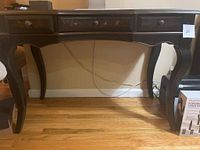 Full view of the small wooden writing desk showing three drawers, ornate drawer knobs and decorative panels, sturdy curved legs, placed against a wall on wooden flooring.