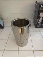 Front view of chrome wine bucket placed on white tile countertop, showing cylindrical shape and polished surface reflecting surroundings.