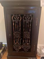 Front view of the black wine cabinet showing decorative ornate metal panels with floral and scroll patterns on the doors.