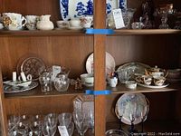 Wooden cabinet shelves filled with various glasses, plates and ceramics including clear glass goblets, hand painted plates, and small decorative items