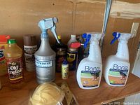 Assortment of cleaning liquids and sprays including Old English oil, Rust-Oleum, WD-40, Bona floor cleaners, with protective gloves and funnel