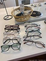 Photo of ten pairs of eyeglasses and sunglasses arranged on a table with some in a golden wire basket.