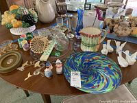 Wide view of assorted home decor items on wooden table featuring platters, pitchers, figurines, glassware.