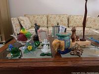 Wide shot of assorted decorative items arranged on a glass table including candle holders, ceramic planter, glass bowl with glass fruits, photo frames, and figurines.