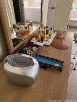 Photo of ultrasonic cleaner, perfume bottles on silver tray, assorted jewelry pieces, and hairbrush on wooden surface with mirror background